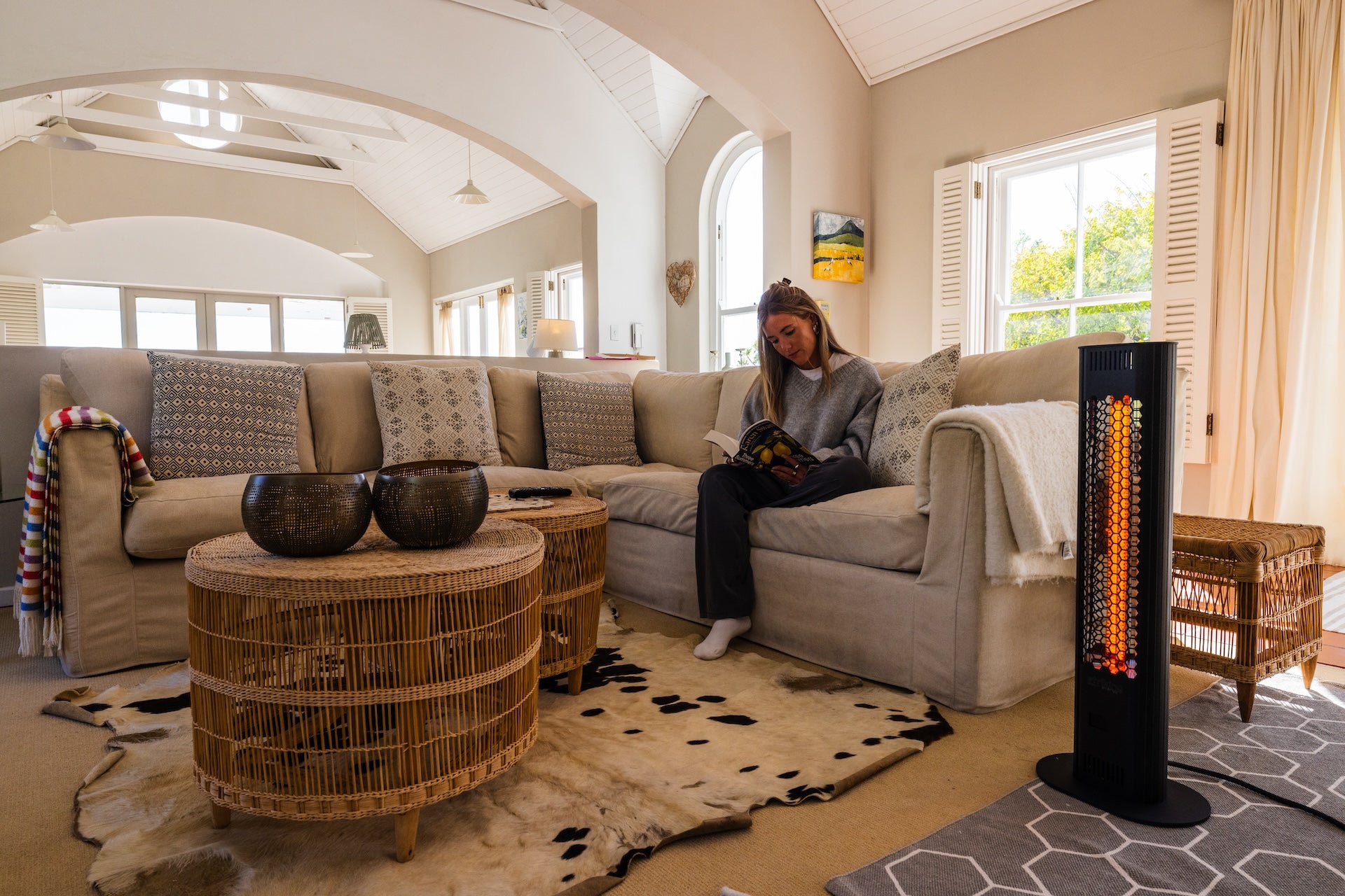 A woman reading comfortably on a modern living room couch next to a tall, freestanding Striking Energy infrared heater emitting a warm glow.