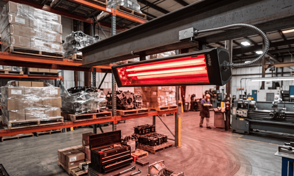 Striking Energy G5 Ruby industrial infrared heater glowing bright red while mounted to an overhead beam inside a busy factory warehouse.