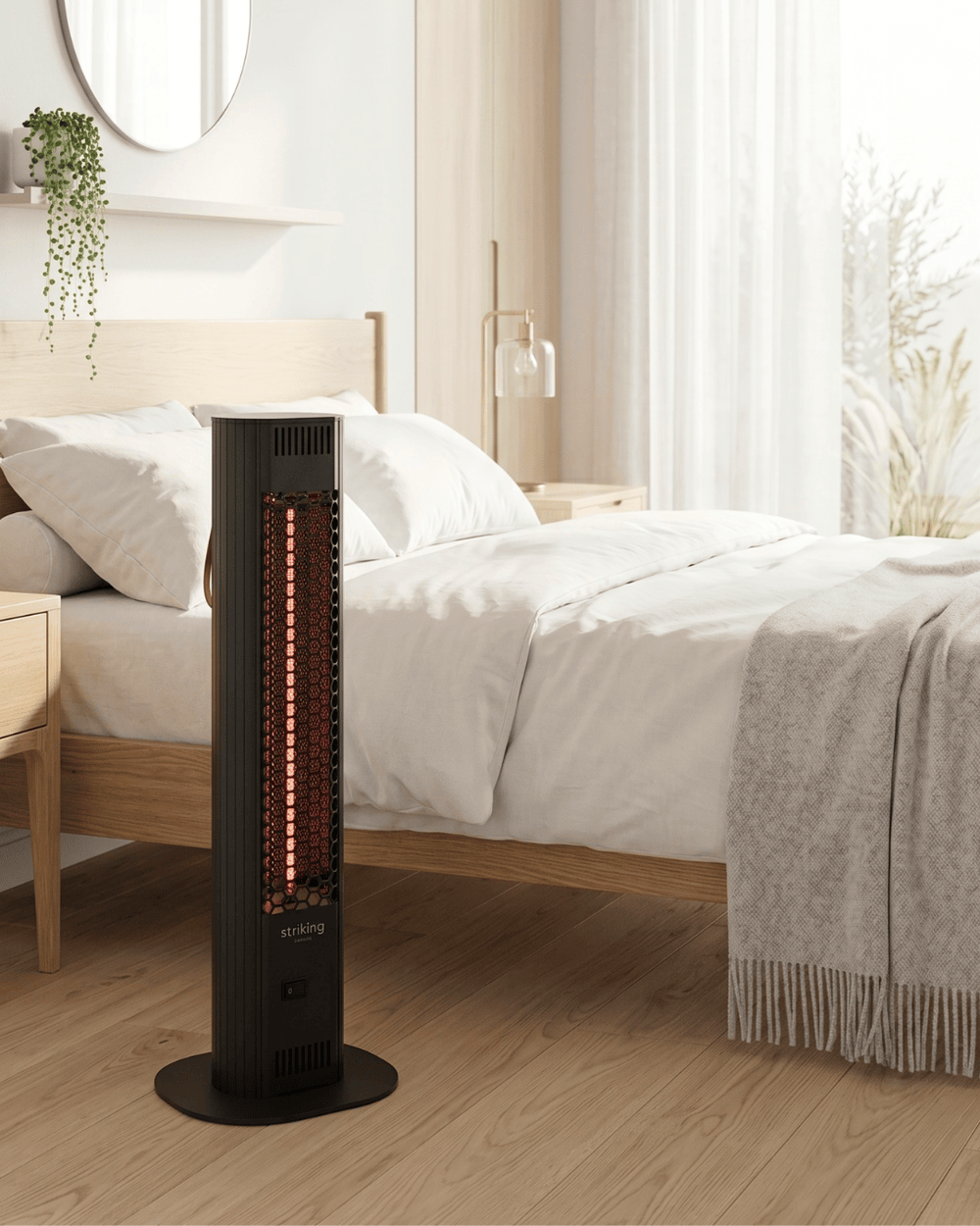 A sleek black Striking Energy freestanding portable infrared heater standing on a wooden floor and glowing warmly next to a modern bed with white sheets.